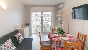 Een knus woongedeelte met een eethoek, een bank met gestreepte kussens en een aan de muur bevestigde televisie.