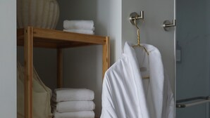 Un baño con un estante de madera con toallas dobladas, una bata blanca colgada en un tozal dorado y una planta en maceta.