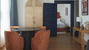 Una habitación con una mesa redonda, dos sillas naranjas, una puerta azul y una cama al fondo.