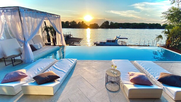 A poolside area with lounge chairs and a table, overlooking a body of water with a sunset in the background. - WTF  HOUSE (Beograd)