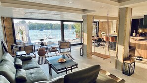 A spacious living area with a large sofa, coffee table, and dining area with chairs. A large window offers a view of the water and boats. - WTF  HOUSE (Beograd)