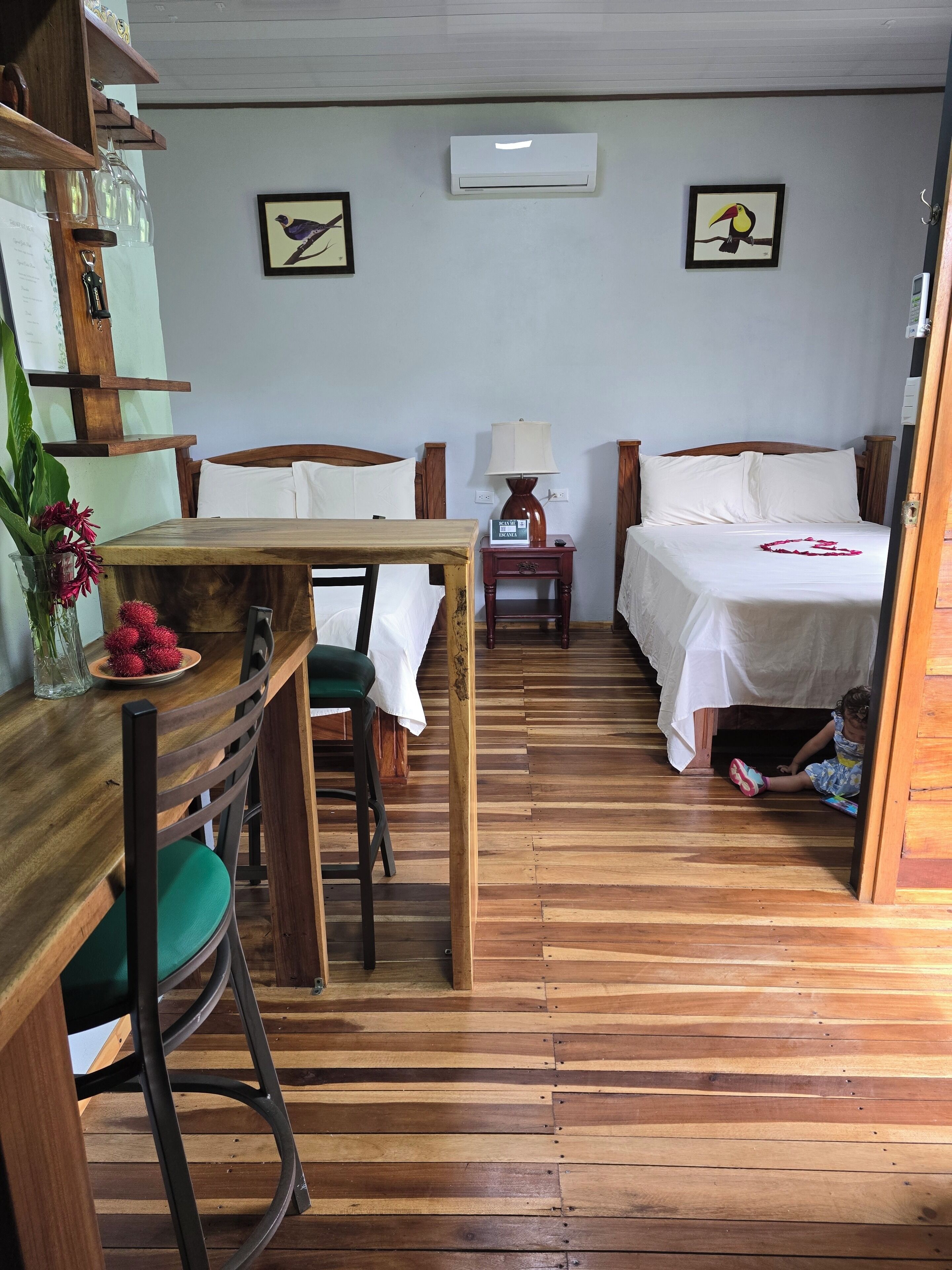 Um quarto com piso de madeira, uma cama com roupa de cama branca, uma mesa com cadeiras, um abajur e duas quadros decorativos na parede.