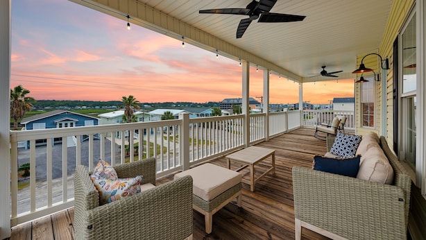 Eine überdachte Veranda mit Korbmöbeln, Deckenventilatoren und Blick auf Häuser und Palmen.