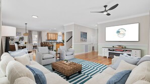 Ein geräumiges Wohnzimmer mit einer großen Couch, gestreiften Sesseln und einem Couchtisch. Ein Fernseher ist an der Wand montiert, und es gibt einen Deckenventilator.