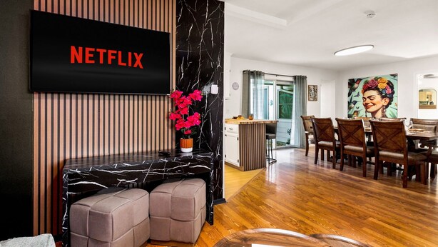 Una sala de estar moderna con un televisor de pantalla plana mostrando 'NETFLIX', una pared de madera con rayas verticales, una mesa con diseño de mármol y un comedor con sillas de madera y un cuadro.