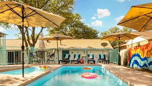 Piscina con sillas de descanso y sombrillas, rodeada por una valla y árboles.
