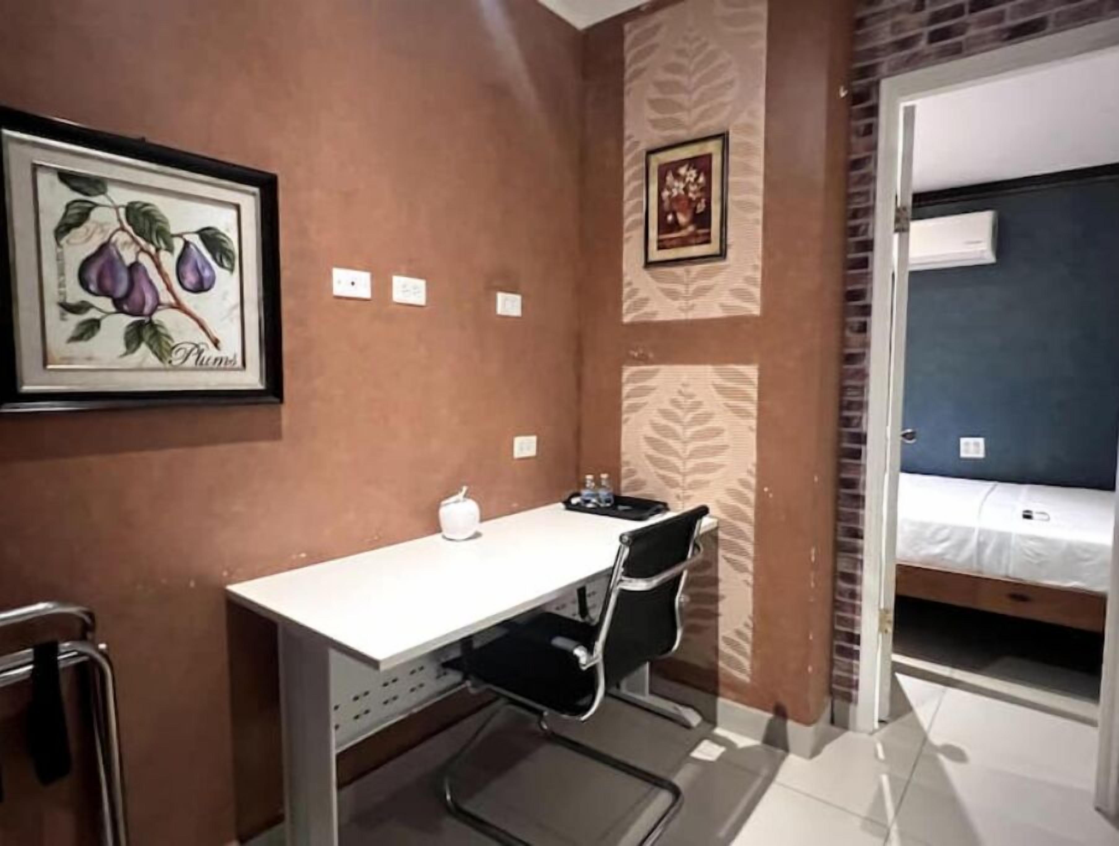 A small, compact room with a desk, chair, and a bed. There are framed pictures on the walls, a water bottle on the desk, and a door leading to another room.