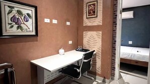 A small, compact room with a desk, chair, and a bed. There are framed pictures on the walls, a water bottle on the desk, and a door leading to another room.