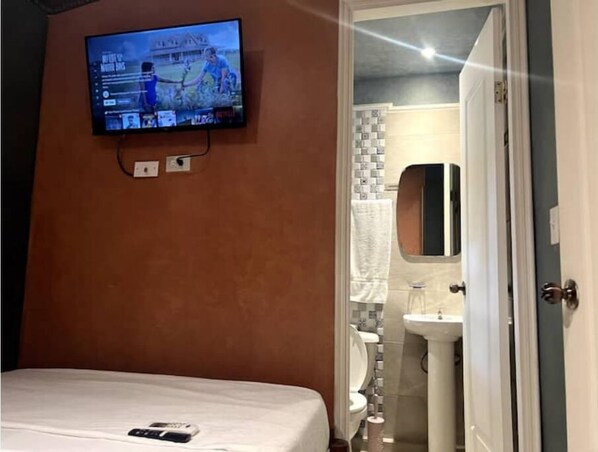 Ein Hotelzimmer mit einem Bett, einem an der Wand befestigten Fernseher und einem durch eine offene Tür einsehbaren Badezimmer.