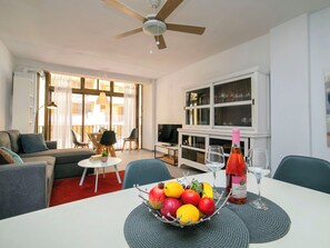 A living room with a sofa, a dining table with a fruit bowl, a TV, and a bookshelf.