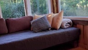 En mysig sittplats med en vadderad bänk, kuddar och en bok på ett träbord.