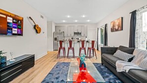 A modern living room with a sofa, a coffee table, and a TV displaying streaming services. There is a kitchen area with cabinets and a dining table with chairs.