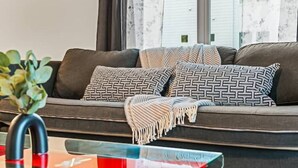 A modern living room with a dark-colored sofa adorned with patterned cushions and a fringed throw blanket. A glass coffee table is in front of the sofa, and a potted plant is visible to the left.