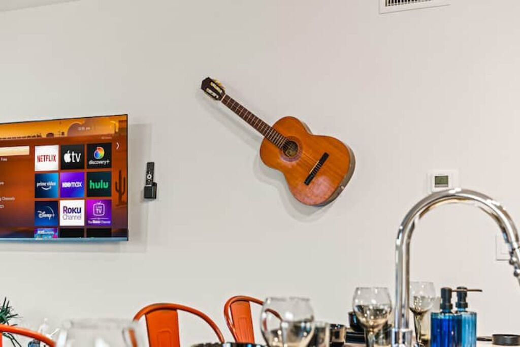 Un téléviseur fixé au mur diffusant différents services de streaming, un ukulélé également fixé au mur, et un évier de cuisine moderne avec de la vaisselle en verre.