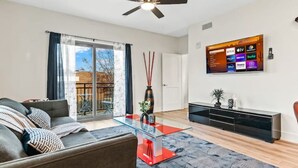 A modern living room with a glass coffee table, grey sofa, and a flat-screen TV mounted on the wall.