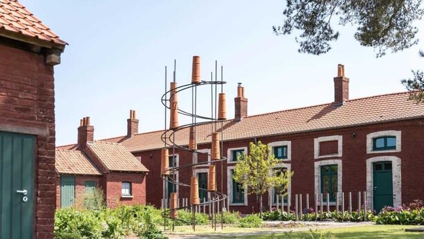 Un bâtiment en briques surmonté d’un toit de tuiles rouges, doté de plusieurs cheminées et agrémenté d’un jardin verdoyant où se trouve une sculpture en métal.