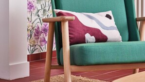 Un fauteuil vert aux pieds en bois et doté d’un coussin à motifs, placé dans une pièce dont le papier peint est à fleurs et un des murs est rayé.