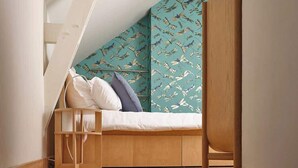 Une chambre mansardée chaleureuse, avec un plafond en pente, un cadre de lit en bois et un papier peint turquoise orné d’un motif d’oiseaux.