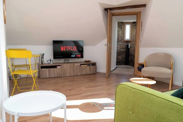 Ein Wohnzimmer mit einem Flachbildfernseher, der 'NETFLIX' anzeigt, einem gelben Klappsessel, einem grünen Sofa und einem kleinen weißen Tisch.
