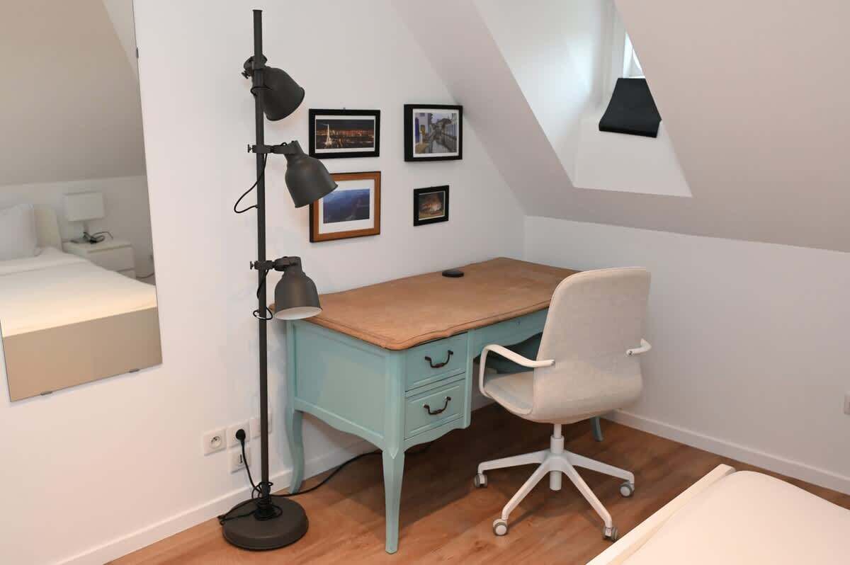 Une petite pièce bien éclairée, avec un bureau, une chaise et un lampadaire. Trois tableaux encadrés ornent le mur, et on aperçoit en arrière-plan une partie d’un lit.