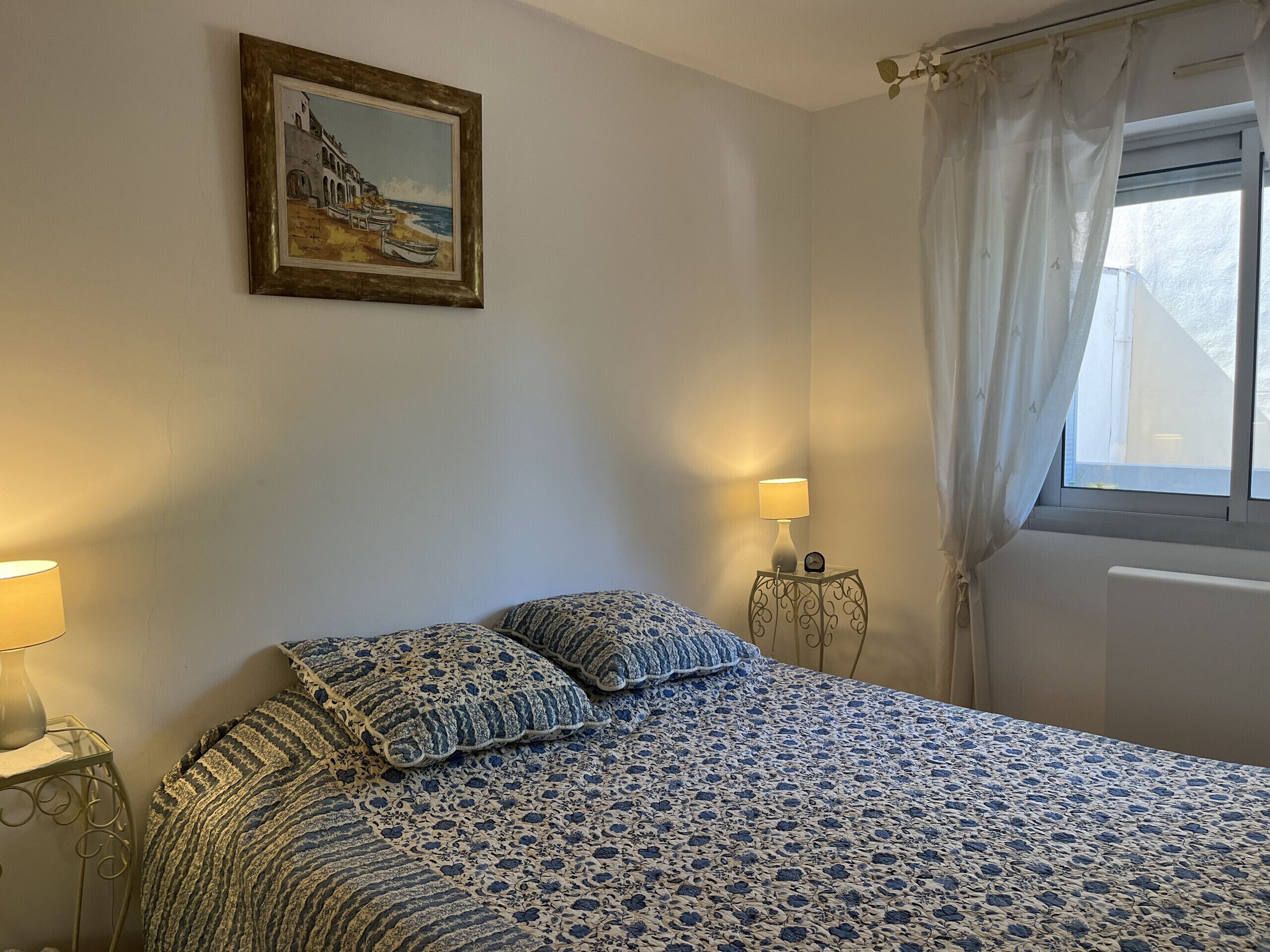A bedroom with a bed, two bedside lamps, a clock, a window with curtains, and a painting on the wall.