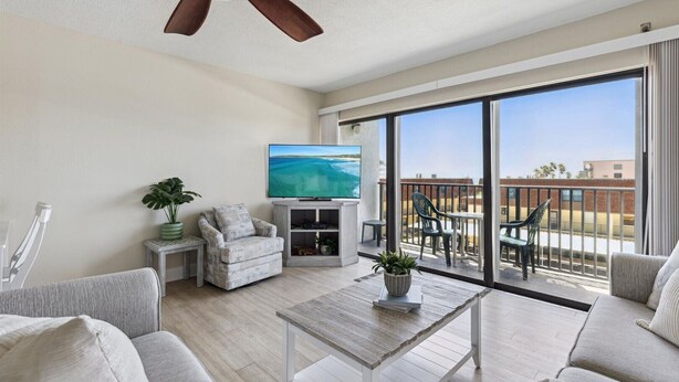 A spacious living room with a ceiling fan, a flat-screen TV, a comfortable sofa, a coffee table, and a balcony with outdoor seating. - Nice and Beautiful Unit - Oceanfront Condo Pool near Attractions (Cocoa Beach)
