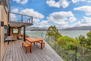 A wooden deck with a table and chairs, overlooking a body of water and mountains.