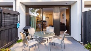 A patio with a round glass table and four chairs, a barbecue grill, and a wooden fence.