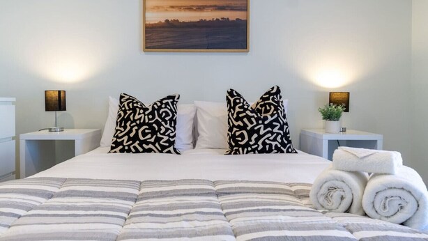 A neatly made bed with striped bedding, two patterned pillows, a framed sunset painting above, and a bedside lamp on each side.