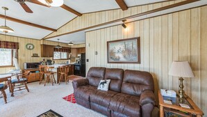 A cozy living room with a brown leather sofa, wooden dining table, and chairs. A kitchen is visible in the background with wooden cabinets and a dining area. - Twice Upon A Lake : Summer Retreat for the Whole Family (Curran)