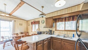 A kitchen with wooden cabinets, a central island, a refrigerator, and a dining area with chairs and a table. - Twice Upon A Lake : Summer Retreat for the Whole Family (Curran)