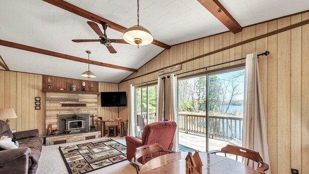 A cozy cabin interior with a fireplace, wooden furniture, and a large window overlooking a deck. - Twice Upon A Lake : Summer Retreat for the Whole Family (Curran)