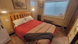 Een slaapkamer met een houten hoofdbord, een groot bed met een rood-beige dekbedovertrek, een raam met jaloezieën, een nachtkastje en een stoel.