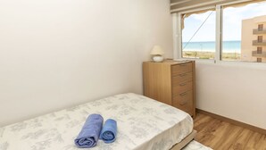 Ein kleines Schlafzimmer mit einem Bett, einem Holznachttisch, einer Lampe und Blick auf den Strand.
