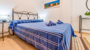 Una cama bien hecha con ropa de cama azul, una mesita de noche con lámpara, y un cuadro en la pared de casas coloridas.