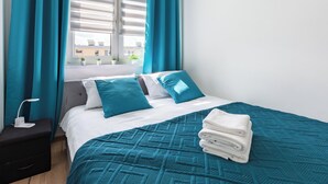 Una cama bien hecha con ropa de cama color aguamarina, una mesita de noche con una lámpara y una ventana con cortinas.