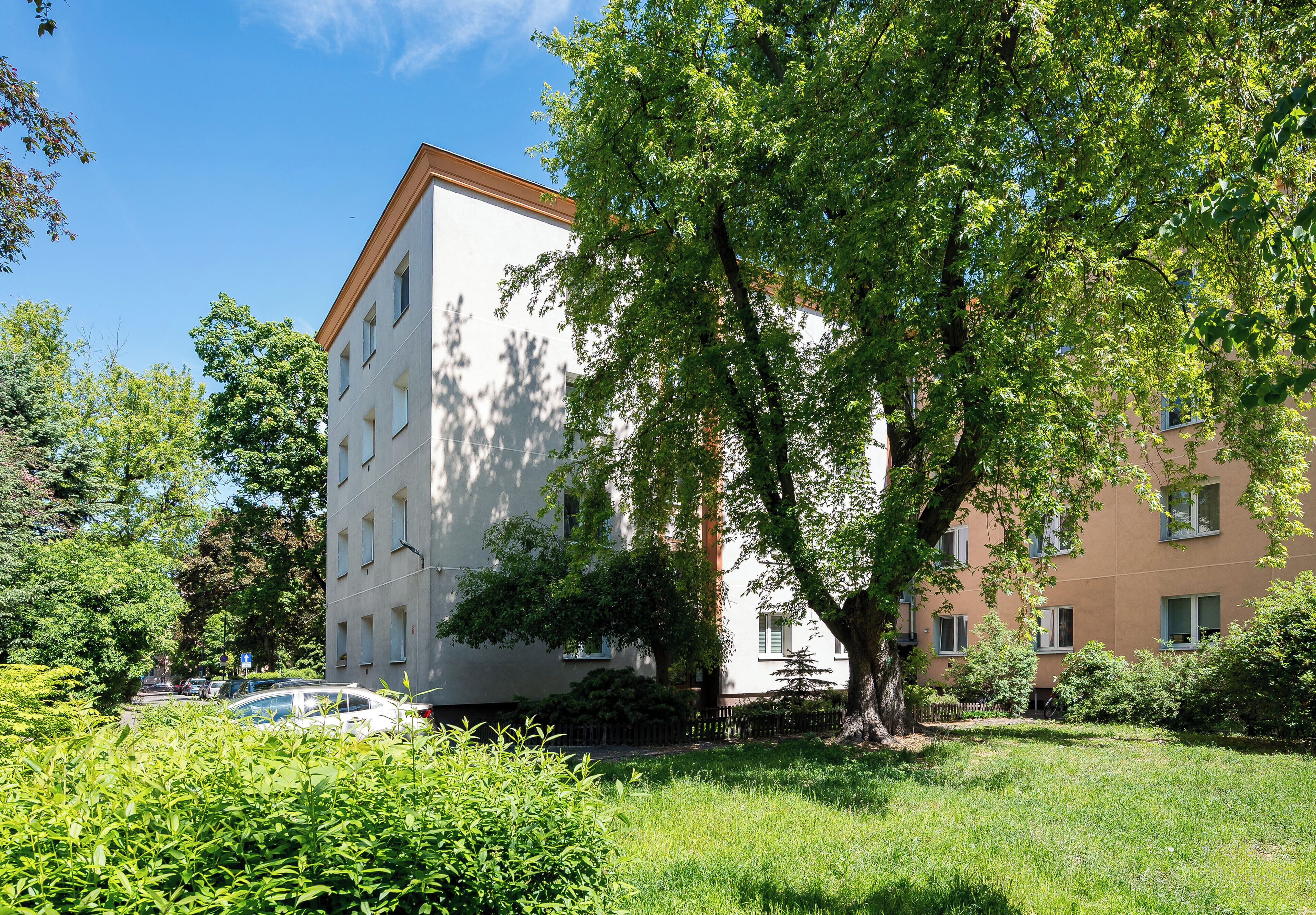 A multi-story building with a white facade and a brown roof, surrounded by lush greenery and trees.