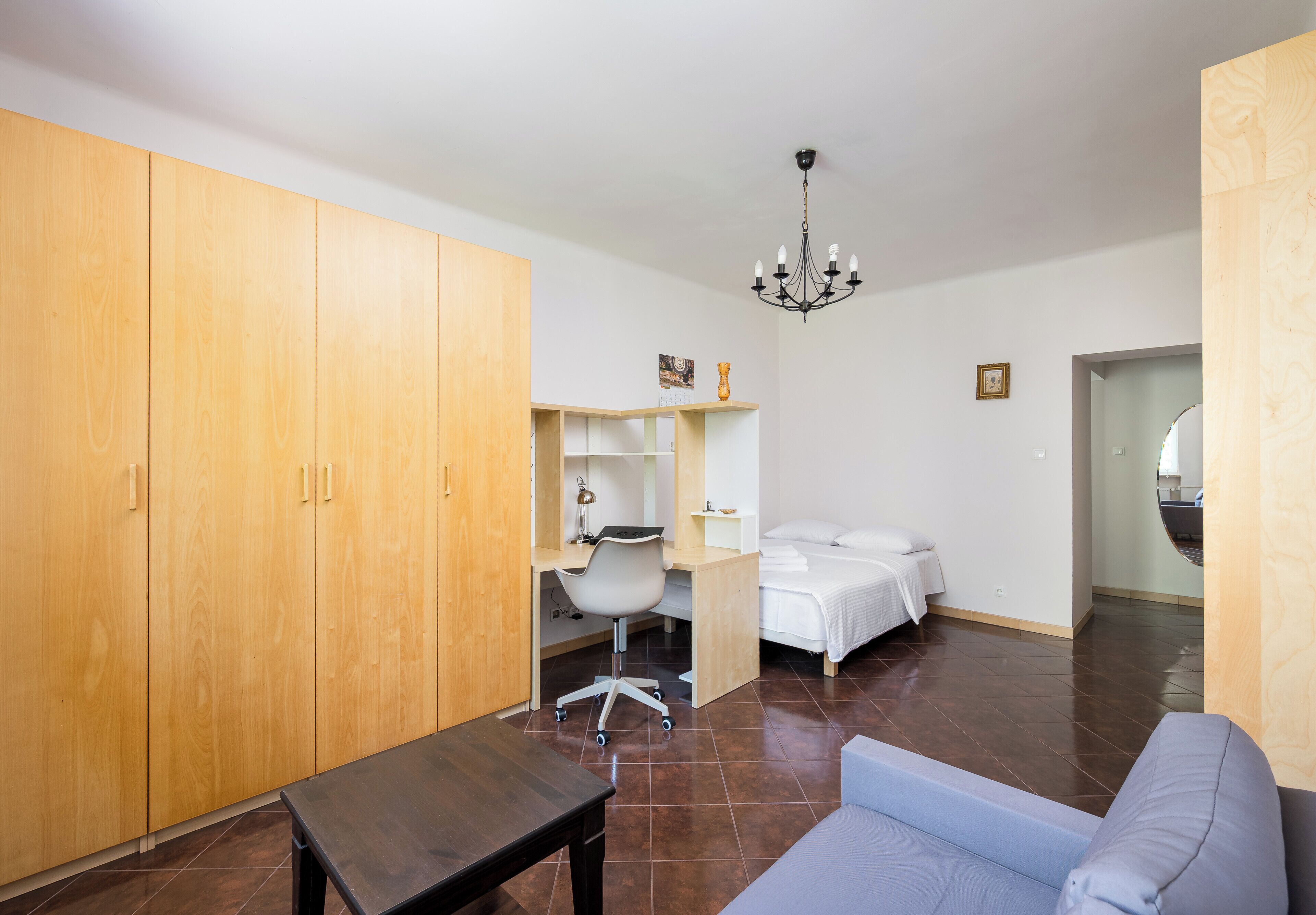 A room with a wooden wardrobe, a desk with a chair, a bed, and a blue sofa.