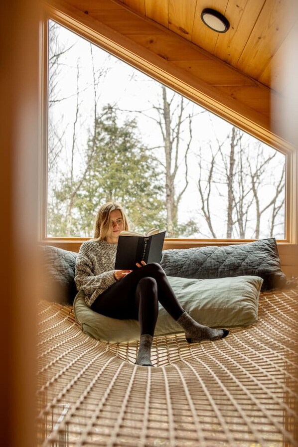 A person sitting on a woven floor mat, reading a book, with a large window and wooden ceiling in the background.