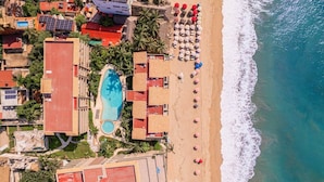 An aerial view of a resort with a swimming pool, beach with umbrellas, and ocean views.
