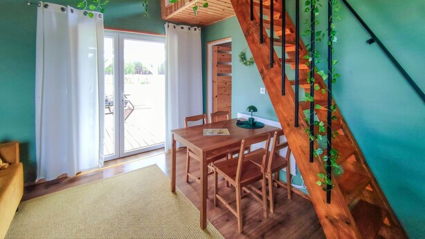 A room with a wooden staircase, a dining table with chairs, a small table with a lamp, and a view through a glass door to an outdoor area.