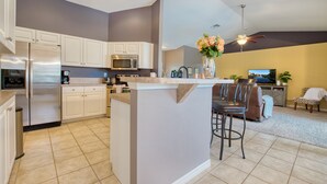 A modern kitchen with white cabinets, a stainless steel refrigerator, a microwave, and a coffee maker. There is a dining area with a bar stool and chairs. - VILLA SALTY ESCAPE - HEATED POOL | FAMILY FRIENDLY | BOAT RENTAL + TOURS VOUCHER INCL. (Cape Coral)