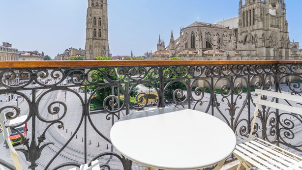 Un balcon doté de balustrades en fer forgé et d’une table blanche, offrant une vue sur une cathédrale historique à deux flèches et sur un clocher.