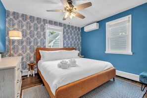 A bedroom with a wooden bed, white bedding, a ceiling fan, patterned wallpaper, a window with blinds, and a small stool.