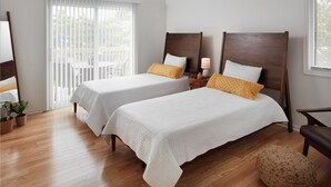 Une chambre d’hôtel avec deux lits, un plancher en bois, un balcon avec des stores blancs et une petite table avec une lampe.
