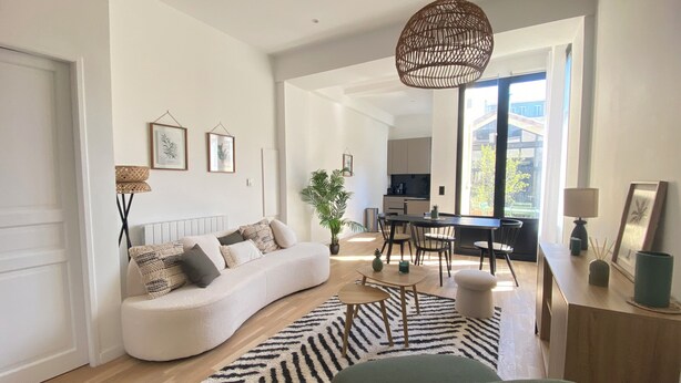 A modern living room with a curved sofa, a dining table with chairs, a large window, and a kitchen in the background.