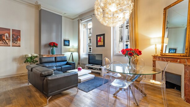 Una sala de estar con sofá, mesa de centro y mesa de comedor de vidrio con sillas. Hay un televisor, una lámpara y una chimenea.