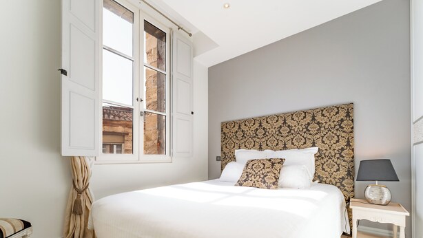 Un dormitorio con una cama grande, una mesita de noche con lámpara, una ventana con cortinas y un cabecero estampado.