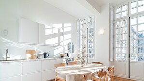 Une cuisine moderne avec des meubles blancs, une table de salle à manger ronde, des chaises en bois et de grandes fenêtres donnant sur des immeubles.