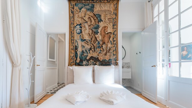 Una cama bien hecha con sábanas y toallas blancas, un tapiz con un jinete y un caballo, y un suelo a cuadros.